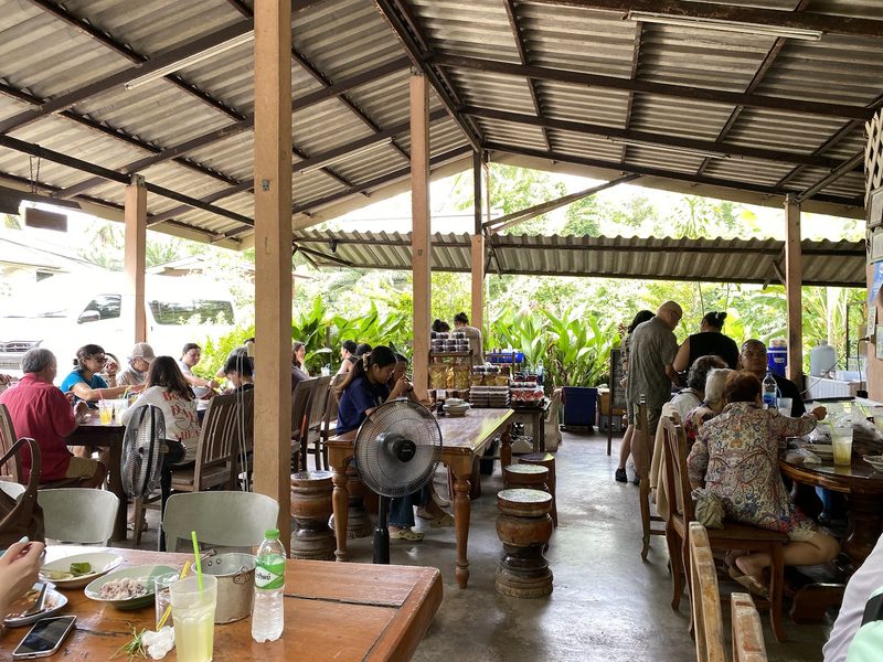บรรยากาศภายในร้านแบบเปิดโล่ง โต๊ะไม้ พัดลม และลูกค้านั่งกินมื้อกลางวัน